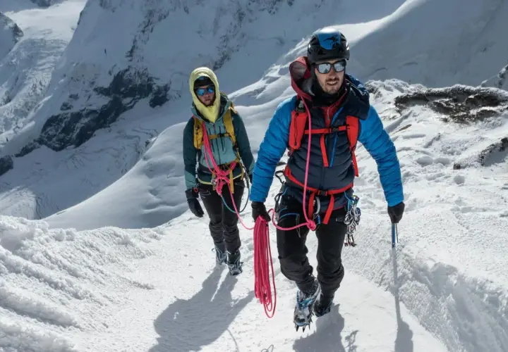 Dwóch alpinistów wspinających się po ośnieżonym zboczu, z liną i sprzętem. Zapewniają sobie asekurację lotną w trudnym terenie.