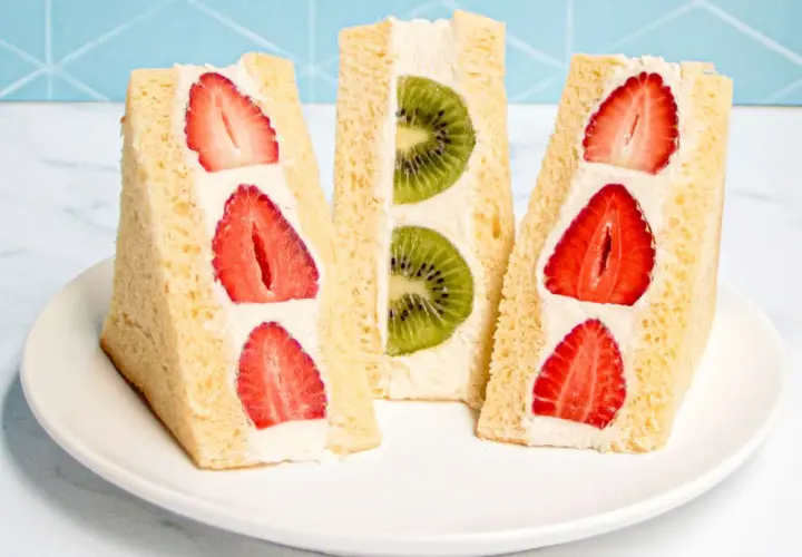 Three triangular Japanese sandwiches filled with fresh strawberries and kiwi slices, nestled in fluffy cream and soft bread.