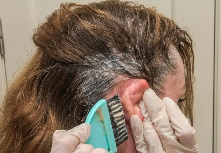 Manos enguantadas usan un cepillo para aplicar tinte en el cabello. Si te manchas la piel, aquí te explicamos como quitar el tinte de la piel.