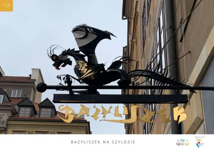 Metalowy bazyliszek ziejący ogniem wisi nad złotym napisem.