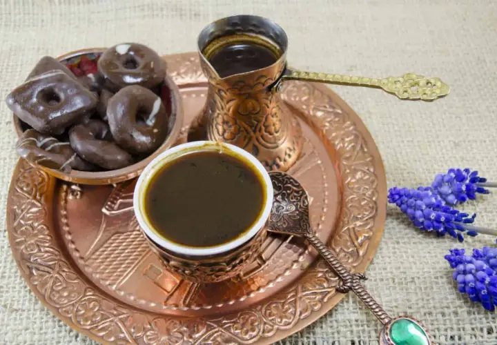 Ein traditionelles Mokkakännchen und eine Tasse mit starkem Mokka-Kaffee, dazu Schokoladenkekse und blaue Blumen auf einem Kupfertablett.