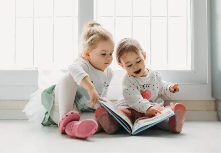 Dwie dziewczynki, jedna 1,5 roczne dziecko mówi tylko po swojemu, przeglądają kolorową książeczkę.