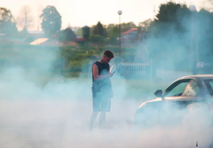 Młody mężczyzna stoi w kłębach dymu, patrząc w telefon. Obok niego samochód, z którego wydobywa się dym, jakby diesel kopcił na czarno.