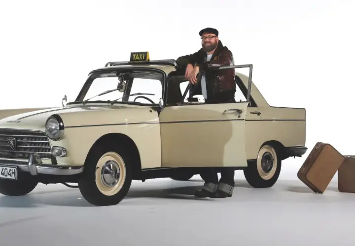 Mężczyzna w skórzanej kurtce opiera się o zabytkowy Peugeot 404 z tabliczką TAXI. Obok stoją walizki.
