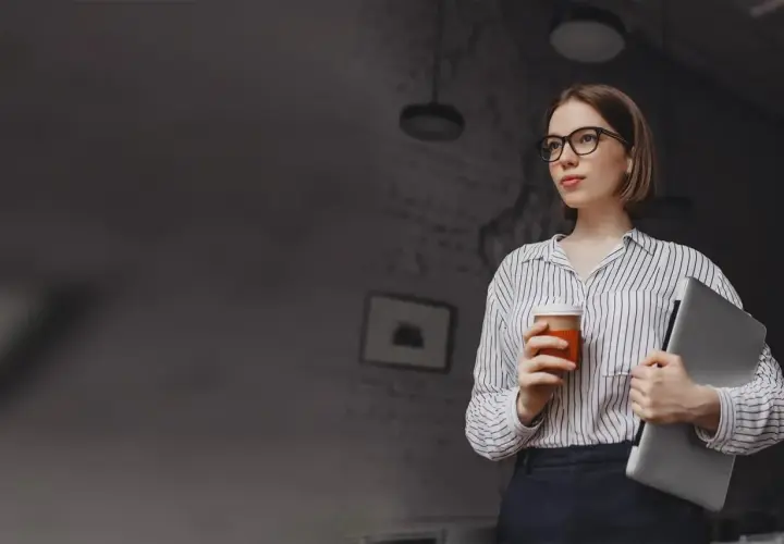 Kobieta w okularach trzymająca kawę i laptopa.