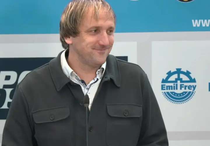 Thomas Neubert lächelt in die Kamera, trägt eine dunkle Jacke und ein gemustertes Hemd. Im Hintergrund sind Logos zu sehen.
