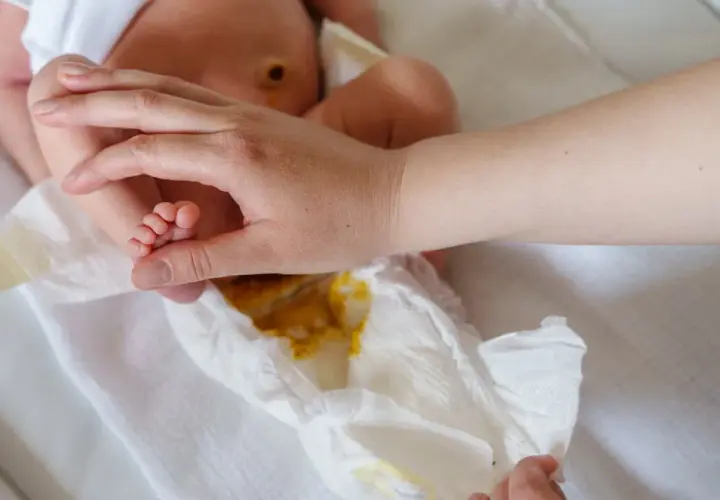Rączka rodzica delikatnie dotyka stópki niemowlaka, podczas gdy pieluszka pokazuje zawartość – kupa niemowlaka.