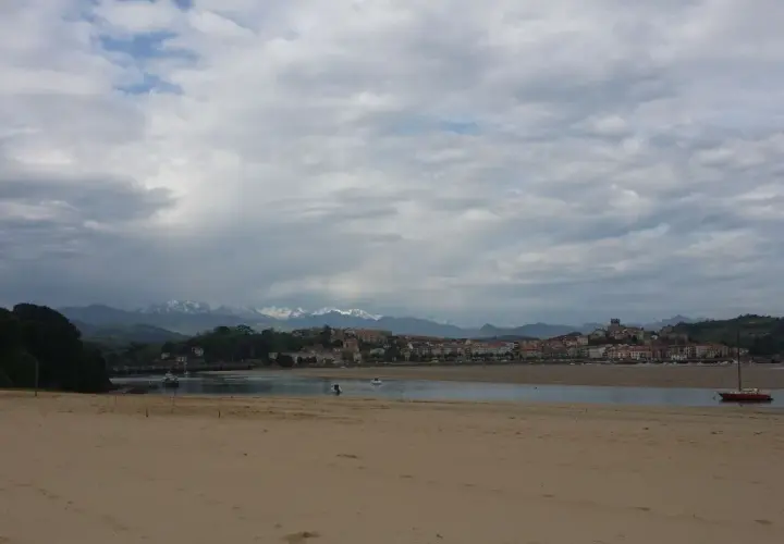Arena dorada en la playa de la Maza, con un velero rojo en el agua y un pueblo costero al fondo, bajo un cielo nublado.