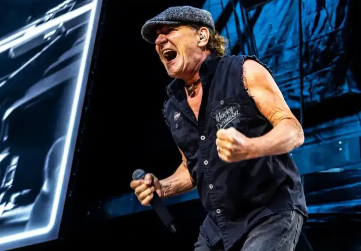 Brian Johnson z zespołu AC/DC na koncercie, w czapce i kamizelce Harley-Davidson, śpiewa z pasją do mikrofonu.