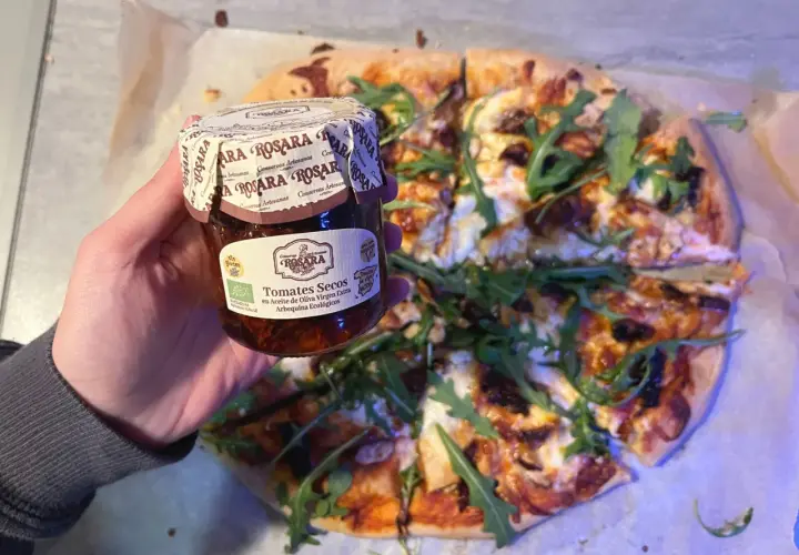 Mano sostiene un tarro de tomates secos en aceite de oliva virgen extra, listo para añadir a una pizza con rúcula.