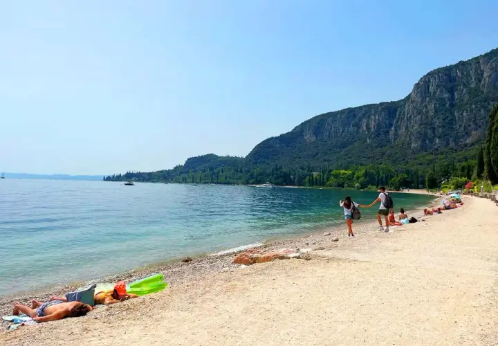 Ein Paar hält Händchen am Strand, während andere am Gardasee schwimmen. Berge und Bäume säumen die Küste.