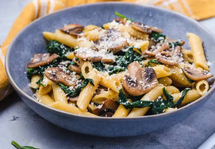 Penne z pieczarkami i szpinakiem, posypane parmezanem, podane w szarej misce.