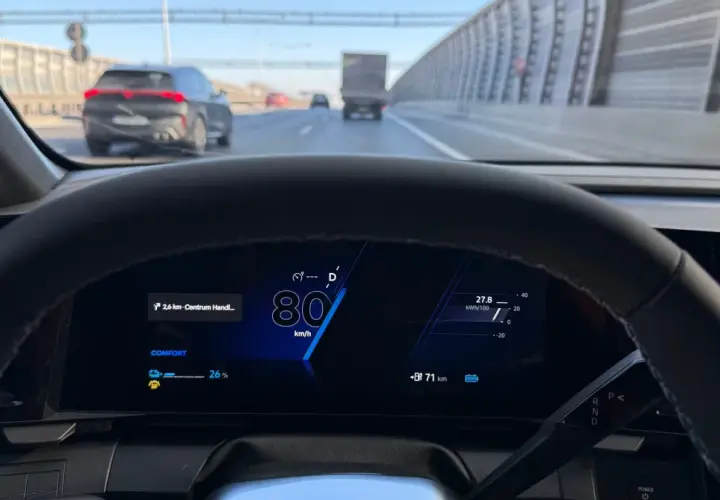 Widok z kabiny samochodu na autostradzie. Na wyświetlaczu 80 km/h, co jest minimalną prędkością na autostradzie. Widać inne pojazdy i barierki.