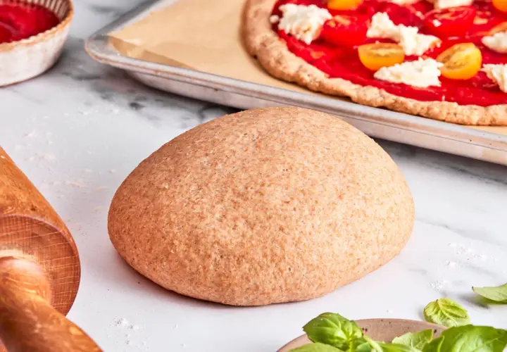 Ein runder Laib vollkorn pizzateig liegt bereit. Im Hintergrund eine fast fertige Pizza mit Tomaten und Mozzarella.