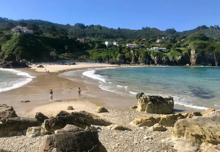 La **playa de Amió** es un paraíso de arena dorada y aguas cristalinas, con colinas verdes y casas blancas al fondo.