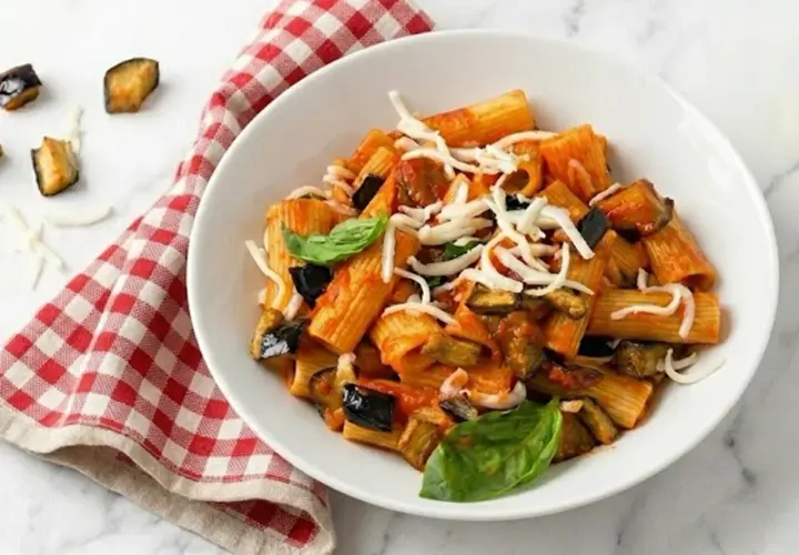 Un plato de pasta alla norma con berenjenas fritas, salsa de tomate y queso rallado, decorado con albahaca fresca.
