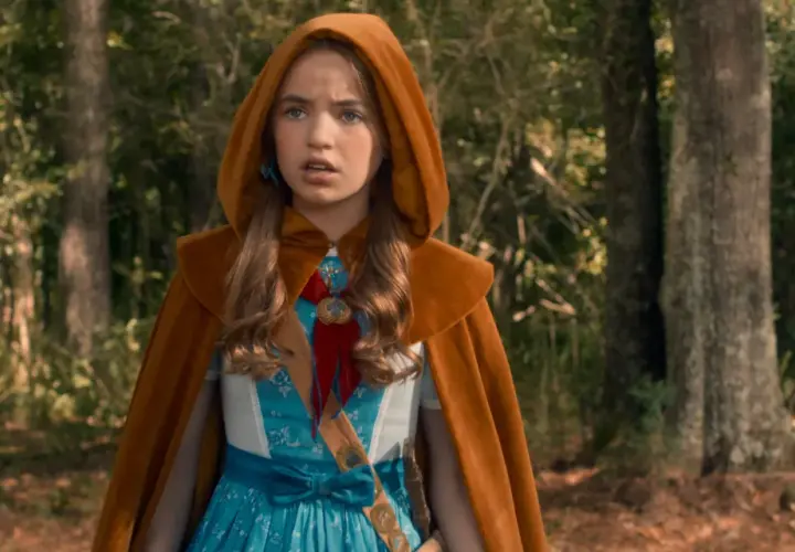 Holly Wheeler z Stranger Things w brązowym płaszczu z kapturem i niebieskiej sukience, stoi w lesie.