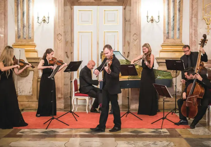 Ensemblen ger en klassisk musik konsert i en elegant sal. Sex musiker spelar fioler, cello och cembalo.