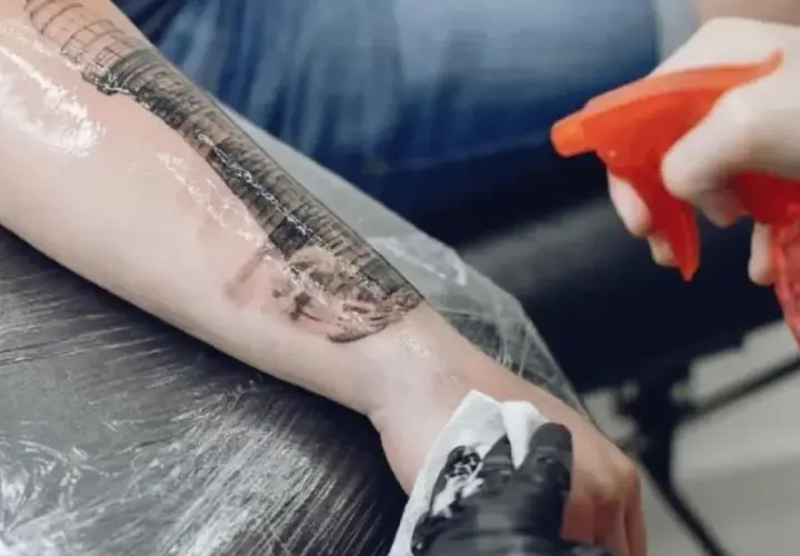 Ręka z rękawiczką aplikuje płyn na świeże tatuaże, które są zabezpieczone folią. To etap pielęgnacji, jak opatrunek second skin.