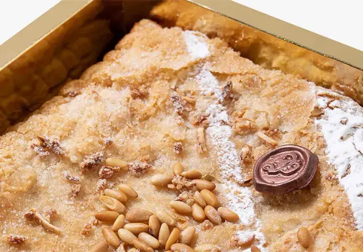 Deliciosa coca de Llavaneres con piñones y un toque de azúcar glas, presentada en una caja dorada.
