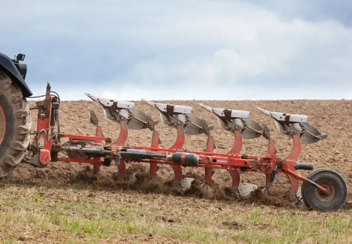 Czerwony pług z wieloma skibami pracuje na polu, przygotowując ziemię do siewu.