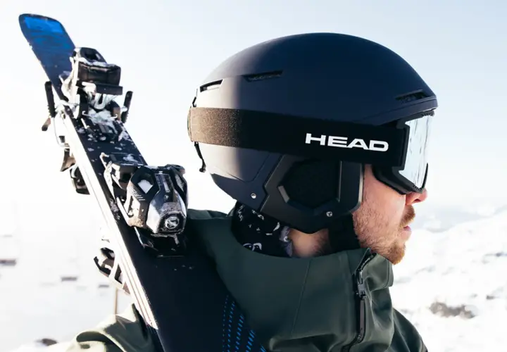 Mężczyzna w kasku narciarskim i goglach HEAD niesie narty. Różnice między kaskiem narciarskim a snowboardowym są subtelne, ale ważne dla bezpieczeństwa.