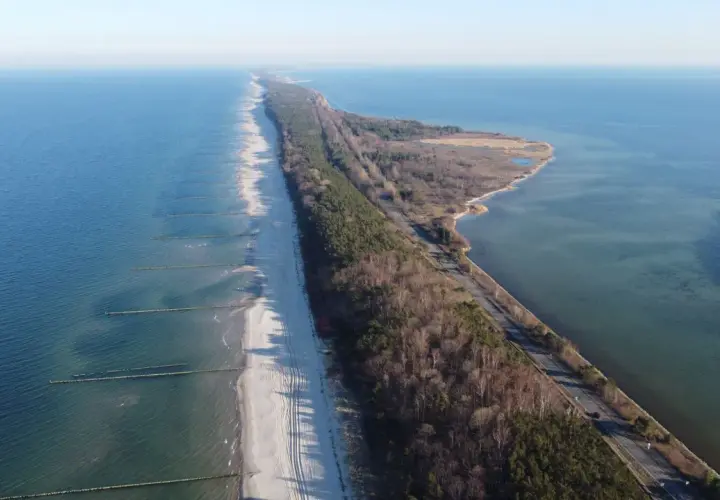 Wąski pas lądu, czyli **półwysep helski**, otoczony przez morze i zatokę. Na plaży widać falochrony, a w głębi las i drogę.