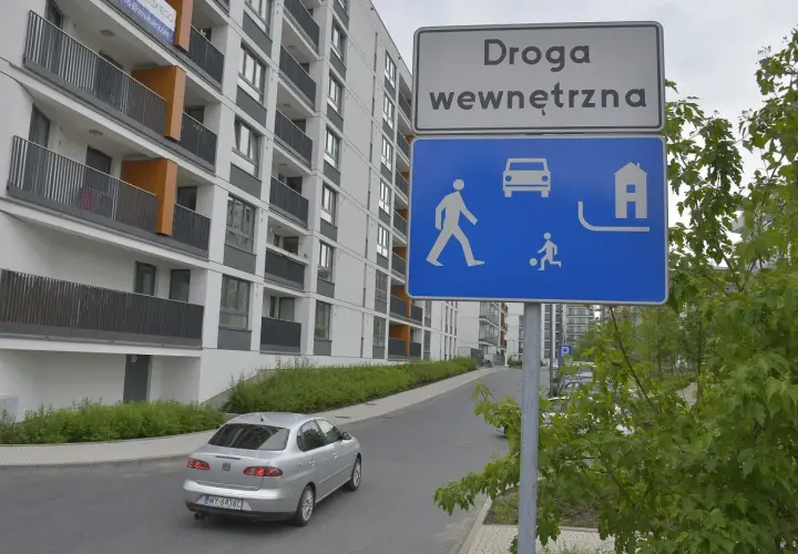 Znak "Droga wewnętrzna" informuje o strefie zamieszkania. Widać tu pieszych, samochód i dom.