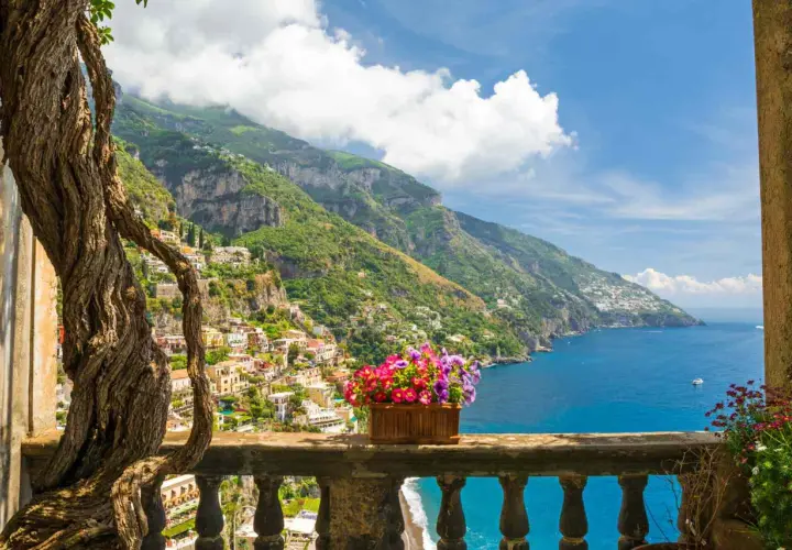 A vibrant view of Amalfi Coast towns clinging to steep green hillsides, overlooking the sparkling blue sea. A twisted tree frames the scene, with a planter of colorful flowers on the balcony railing.
