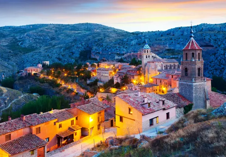 Pueblo español iluminado al anochecer, ideal para escapadas fin de semana España. Casas con tejados de teja y torres de iglesias se alzan entre colinas.