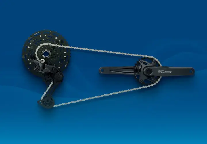 Napęd rowerowy Shimano CUES: korba, przerzutka i kaseta połączone łańcuchem na niebieskim tle.