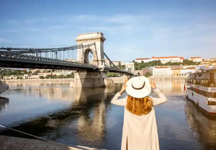 Kobieta w kapeluszu podziwia Most Łańcuchowy w Budapeszcie. W tle widok na Dunaj i budynki, a w portfelu czeka budapeszt waluta na dalsze zwiedzanie.