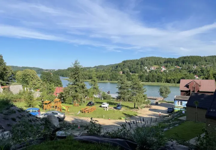 Widok na jezioro i ostrzycki las. Na pierwszym planie plac zabaw i samochody, w tle domki i zielone wzgórza.