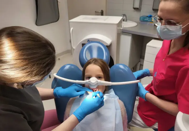 Dziecko u dentysty z maską do podawania gazu rozweselającego. Dwie higienistki dbają o komfort małego pacjenta podczas zabiegu.