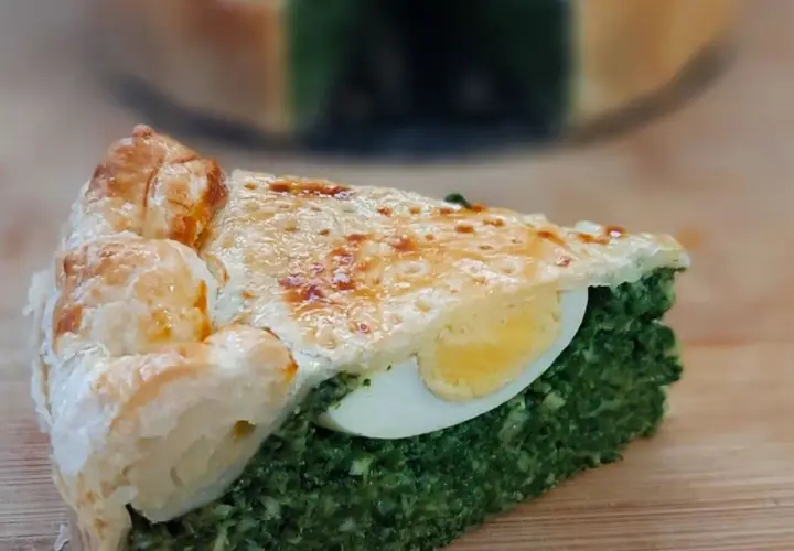 Kawałek torta pasqualina z zielonym nadzieniem i jajkiem, na drewnianej desce.