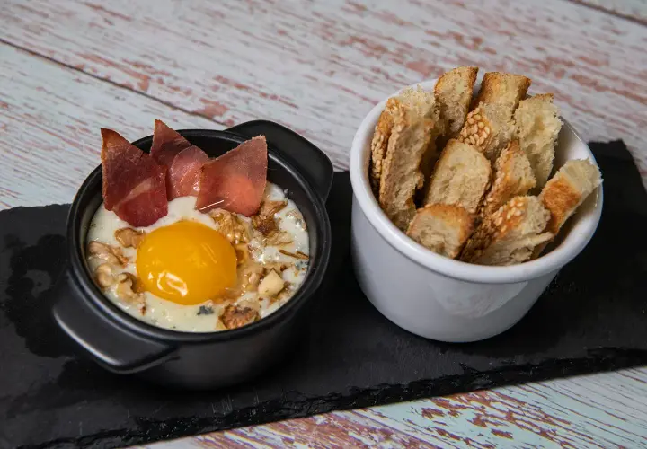 Un œuf cocotte raclette, crémeux et gourmand, garni de noix et de fines tranches de viande séchée, servi avec des toasts croustillants.