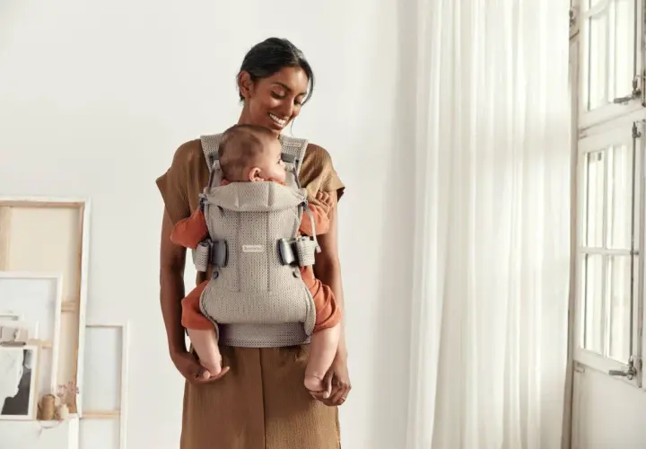 Uśmiechnięta mama tuli dziecko w nosidełku Babybjorn. Od kiedy można używać? Od pierwszych dni życia!