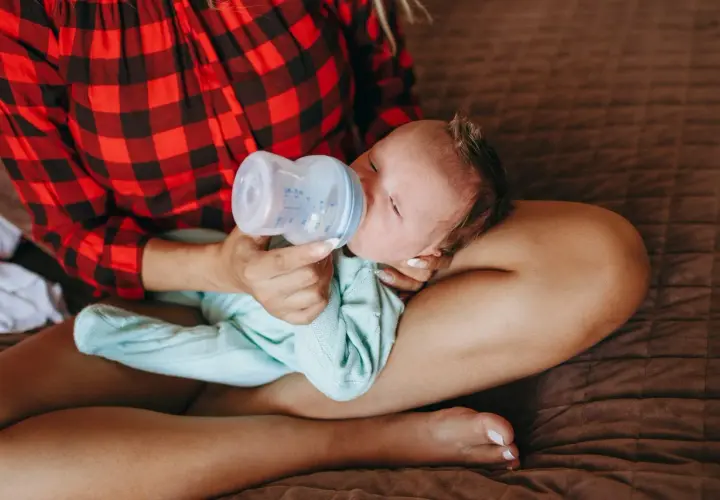 Mamma con maglia a quadri rossi offre il biberon a un neonato, praticando l'allattamento misto.