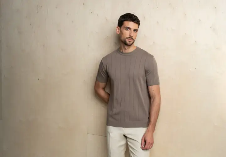 Mężczyzna prezentuje elegancki, casualowy styl. Brązowa, prążkowana koszulka z krótkim rękawem i beżowe spodnie tworzą harmonijną całość.