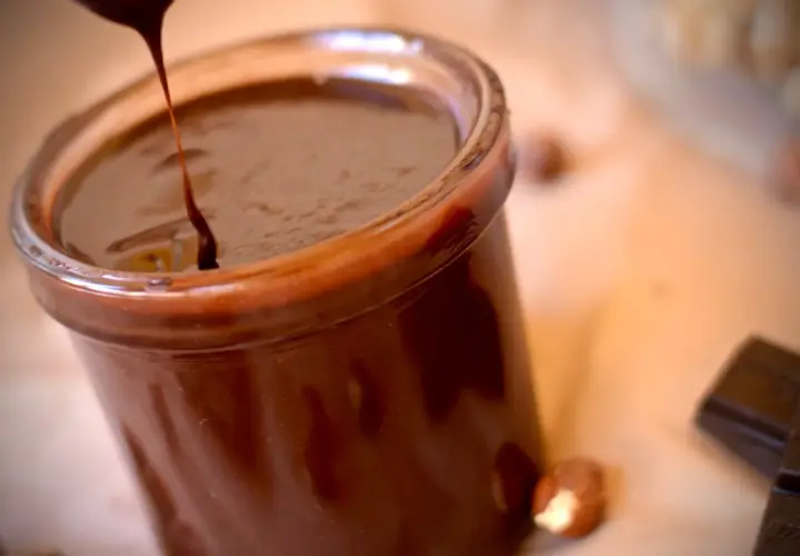 Un chorrito de crema de chocolate cae en un frasco lleno de esta delicia. Avellanas y tabletas de chocolate completan la escena.