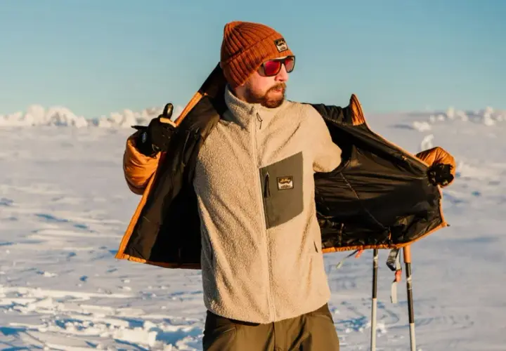 Man i orange mössa och solglasögon visar upp sin lager på lager-klädsel i snölandskap med skidstavar.