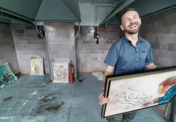 Uśmiechnięty kossakowski malarz prezentuje swoje dzieła. W tle widać obrazy i gaśnicę.