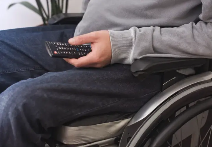 Osoba na wózku inwalidzkim, z umiarkowanym stopniem niepełnosprawności, trzyma pilot do telewizora.