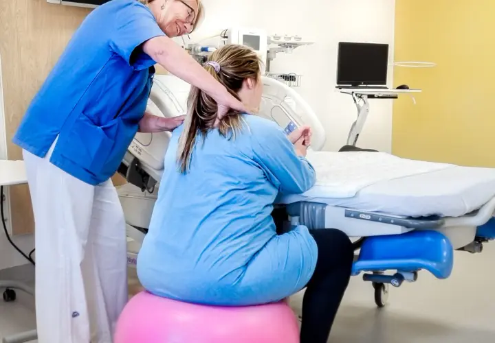 Barnmorska stöttar en kvinna på en rosa boll, v 34. De är i ett förlossningsrum med medicinsk utrustning.