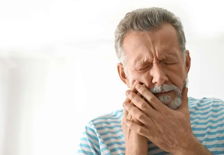 Hombre mayor con los ojos cerrados y la mano en la mejilla, sufriendo de dolor, posiblemente por alveolitis seca.