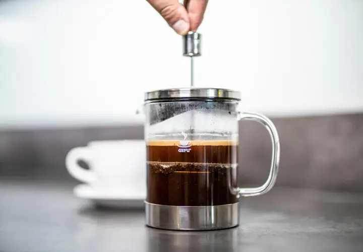 Hand drückt Kolben in French Press, um einen milden Kaffee zuzubereiten. Eine weiße Tasse steht im Hintergrund.