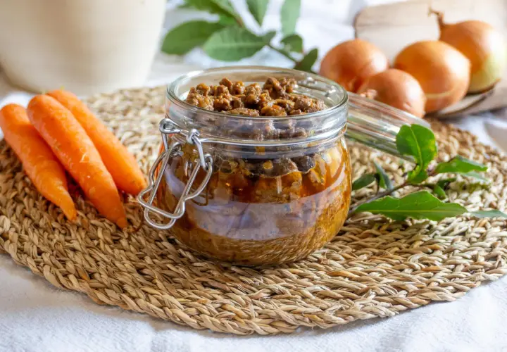 Un vasetto di vetro pieno di saporito ragù di anatra, con carote e cipolle sullo sfondo.