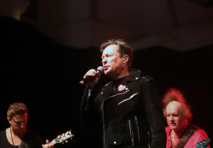 Kawalec śpiewa, a budka suflera czuwa. Muzycy na scenie, w tle gitara i czerwona koszula.