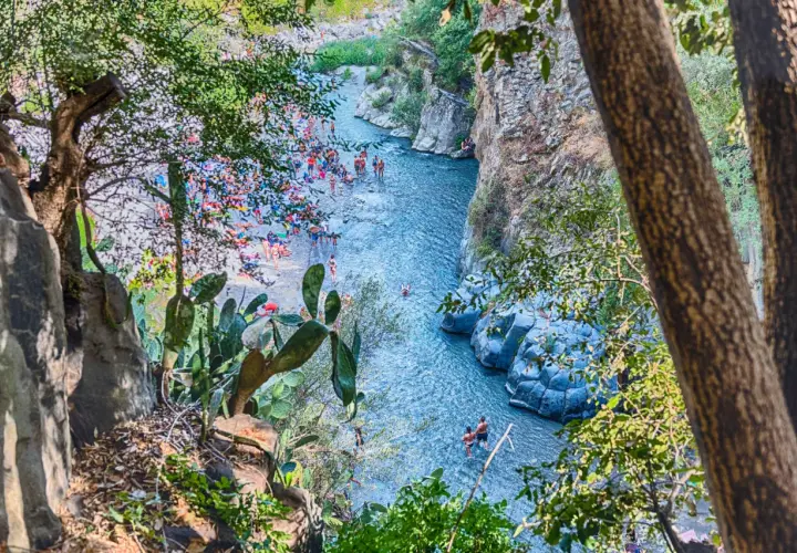 Gente si gode il sole e l'acqua cristallina nella Valle dell'Alcantara, tra rocce vulcaniche e vegetazione rigogliosa.