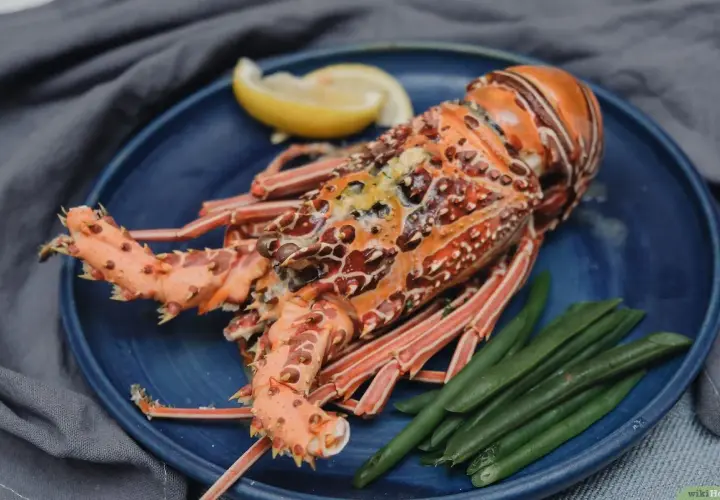 Una langosta marisco, cocinada y servida con judías verdes y una rodaja de limón en un plato azul.